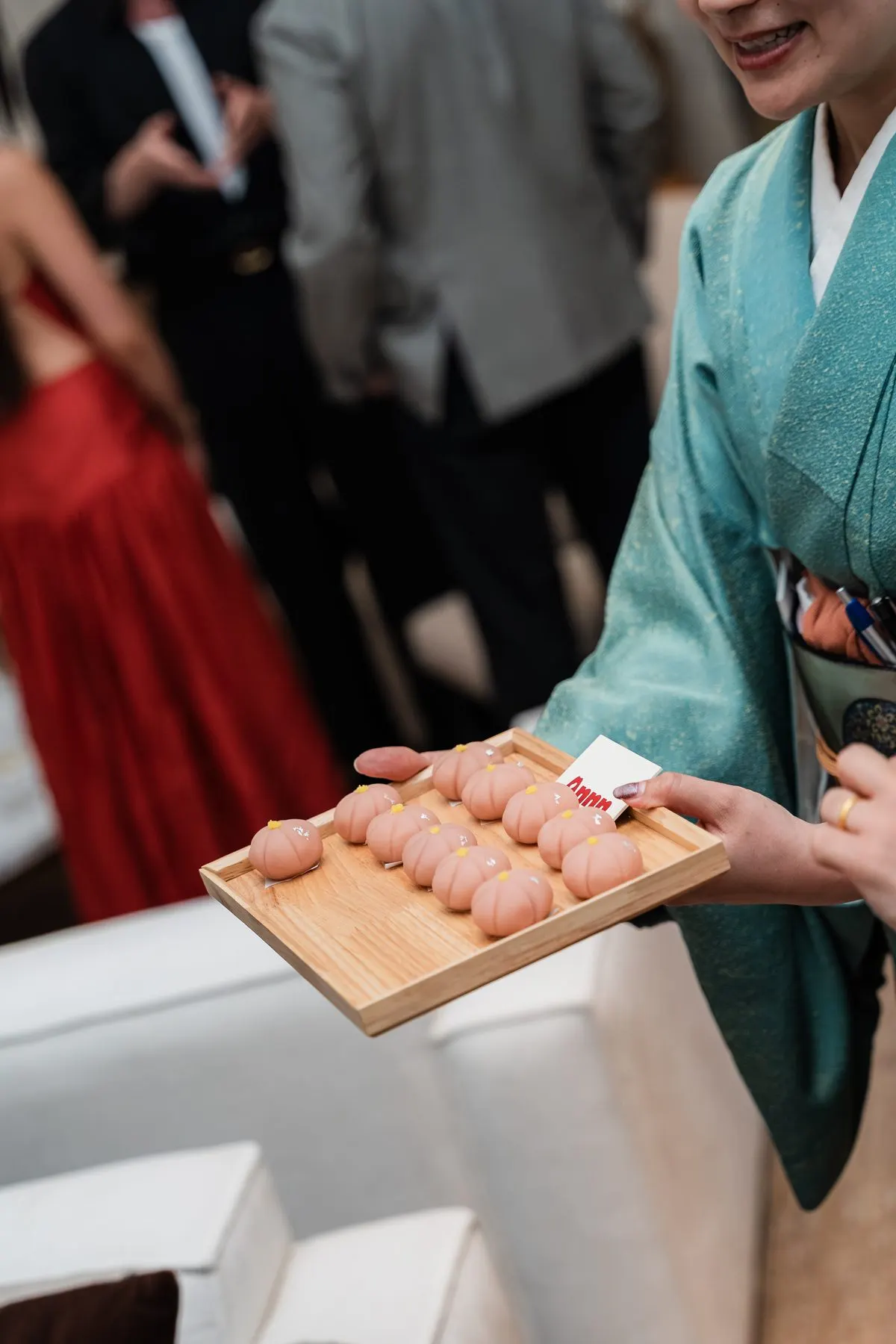 A host in teal kimono offering a tray of pink wagashi